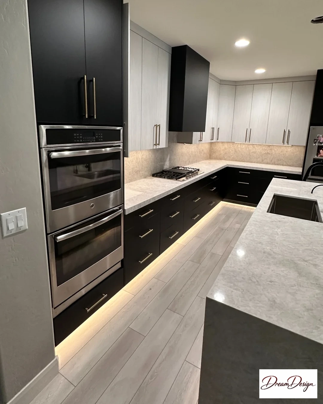 Black and pale kitchen cabinetry with stacked ovens, a sink island and illuminated cabinet bases.