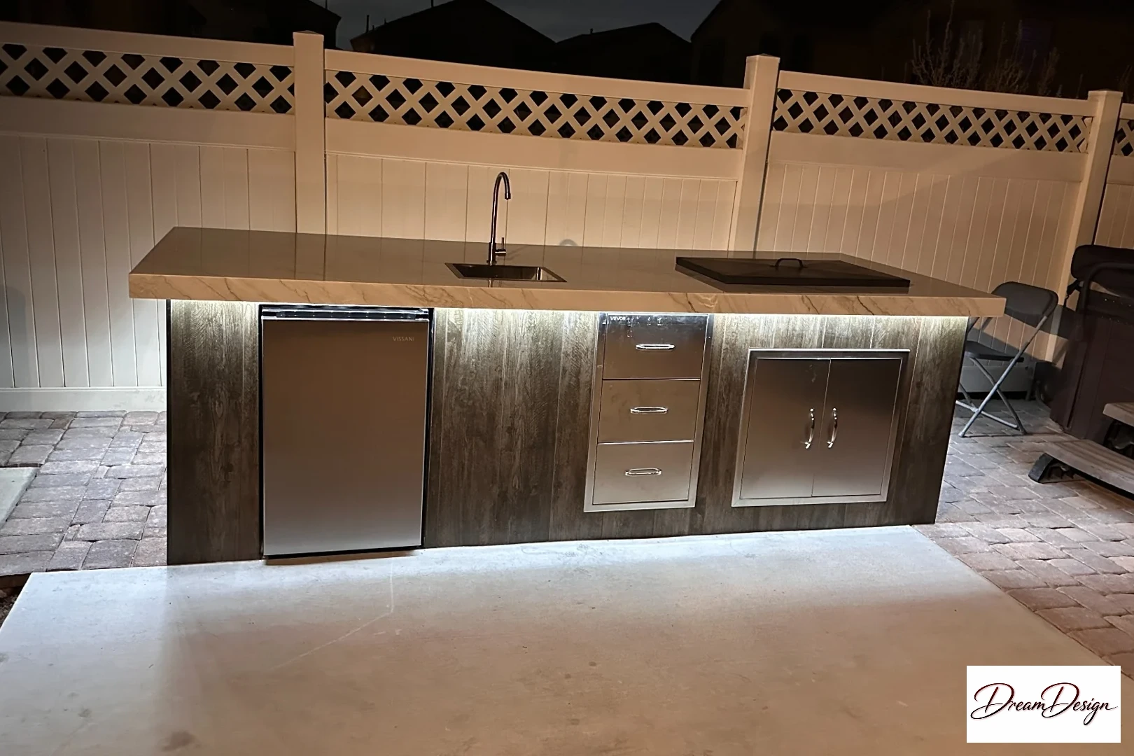 Outdoor counter with a sink, undercounter refrigerator and metal-front storage at night.