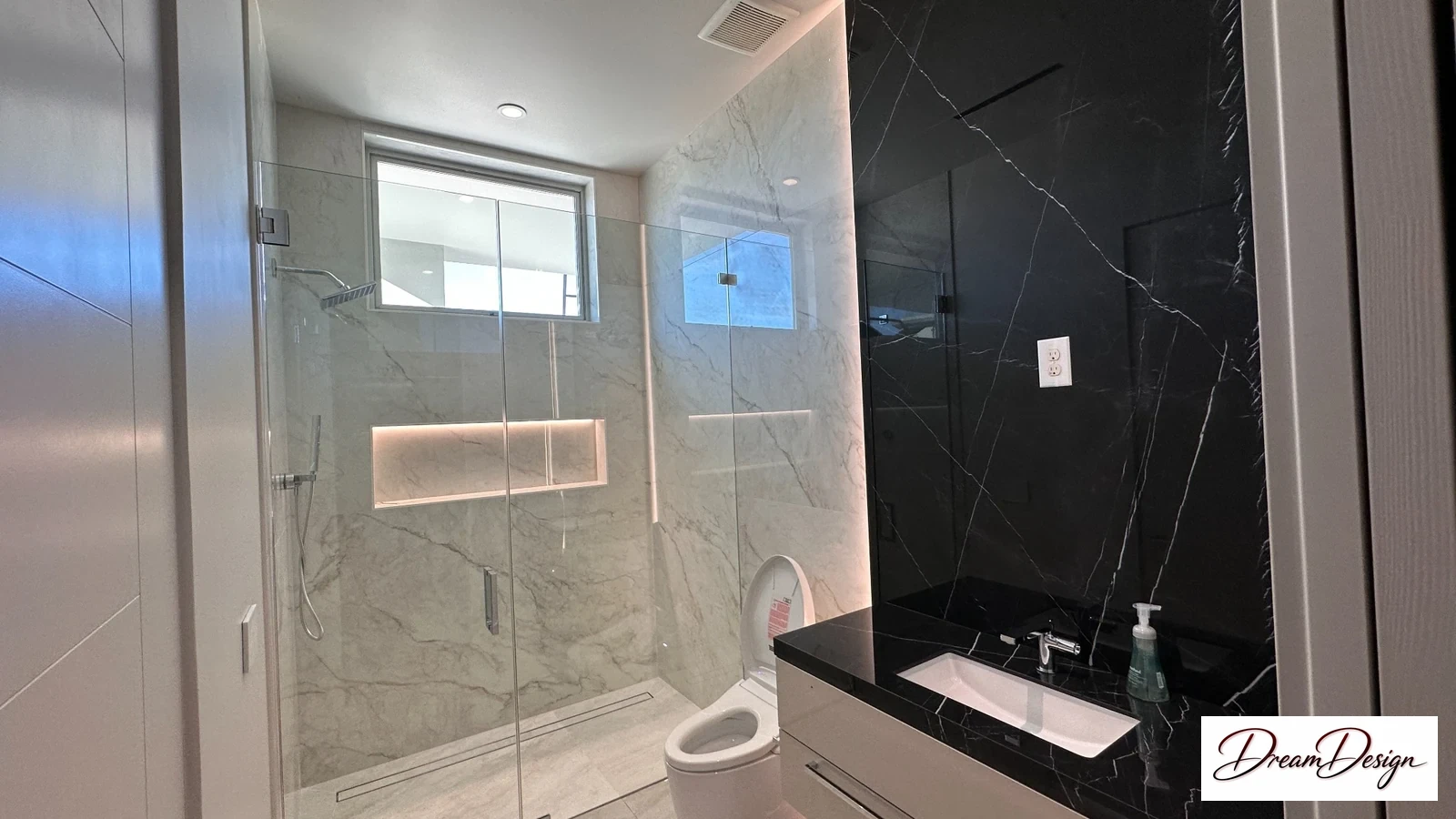 Glass shower enclosure with light veined tile, a window and a dark vanity alongside.