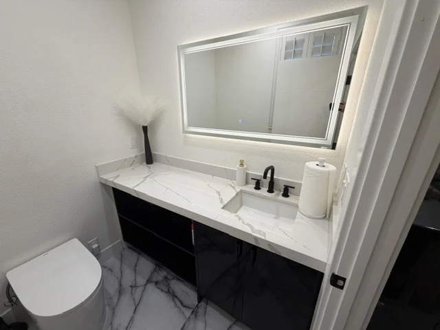 Compact bathroom with a black single-sink vanity, pale veined counter and illuminated rectangular mirror.