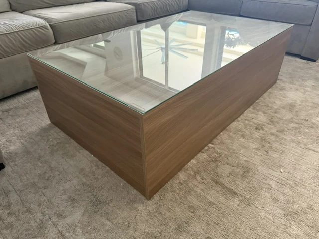 Angled view of a rectangular wood-grain coffee table with a glass top beside a sofa.