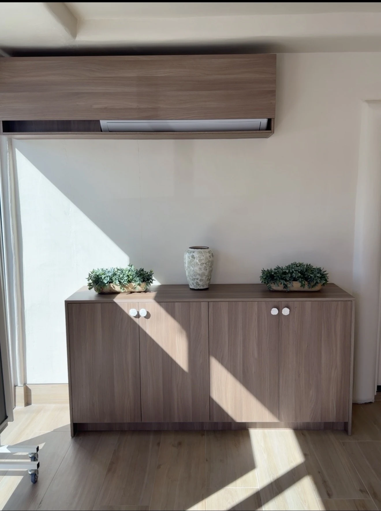 Wood-grain sideboard with round white knobs and matching overhead enclosure in a sunlit room.