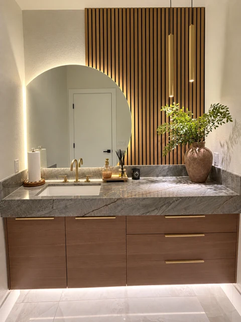 Floating wood-grain vanity, veined countertop, round backlit mirror and vertical wood-tone slat wall.