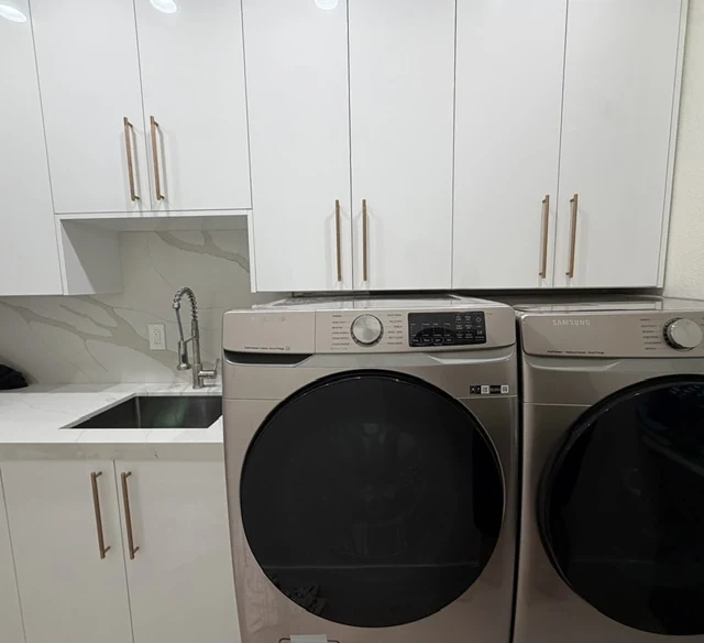 White laundry cabinets with gold-tone pulls, a sink and a light veined backsplash beside two appliances.
