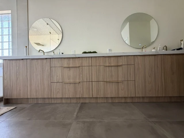 Long wood-grain double vanity with a pale counter, gold-tone pulls and two round mirrors.